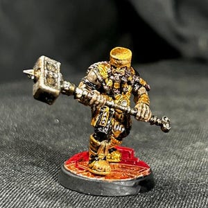 Può includere: Una statuetta in miniatura dipinta di un guerriero nano che tiene un martello. Il nano indossa un'armatura dorata e un elmo. La miniatura è in piedi su una base nera con un design rosso e dorato.