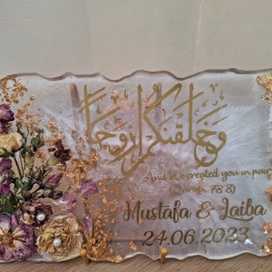 May include: A white resin plaque with gold leaf accents and dried flowers. The plaque features Arabic calligraphy and the text "And We created you in pairs (Quran 78:8)" and "Mustafa & Laila 24.06.2023".