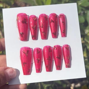 May include: A set of press-on nails in a vibrant pink hue, adorned with gold leaf accents. The nails are displayed on a white card, showcasing a glossy finish. The design features a rectangular shape, perfect for a manicure.