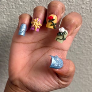 May include: A hand with long, square-shaped acrylic nails. Each nail features a different design, including a blue glitter nail, a yellow flower, a red rose, and a white flower with green leaves. The nails are painted with bright colors.