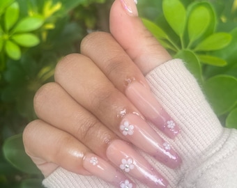 May include: Long, nude-colored acrylic nails with a gradient of pink at the tips. Each nail is adorned with a small, white flower accent.