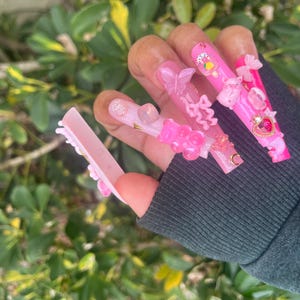 May include: Pink and white long acrylic nails with 3D embellishments, including butterflies, hearts, and bows. The nails are decorated with glitter and gold accents.
