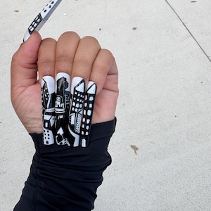 Könnte beinhalten: Eine Hand mit langen, weißen und schwarzen Acrylnägeln. Die Nägel zeigen ein Stadtbild-Design mit schwarzen Gebäuden und weißen Akzenten. Der Daumennagel ist abgelöst und wird von den Fingern gehalten. Die Person trägt ein schwarzes Langarm-Oberteil.