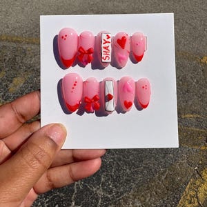 May include: A set of pink artificial nails with red tips, bows, and heart designs. Some nails feature the name "SHAY" and an envelope design. The nails are displayed on a white card.
