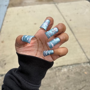 May include: A hand with long, square-shaped artificial nails. The nails are painted with a light blue base and a pattern of pink and white swirls. One nail features a small silver crescent moon. The person is wearing a black long-sleeved shirt.