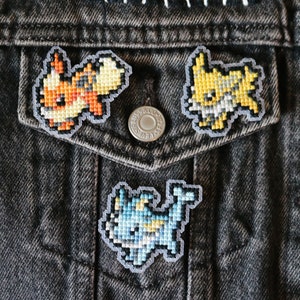 May include: Three embroidered patches featuring Pokemon characters on a dark denim jacket. The patches are of a blue and yellow water-type Pokemon, a yellow electric-type Pokemon, and an orange and yellow fire-type Pokemon.