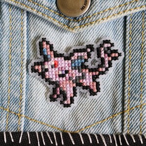 May include: A pixelated patch featuring a pink and black creature with blue accents, sewn onto a light blue denim jacket pocket. The patch has a black border and is stitched with various colors. The denim jacket has yellow stitching.