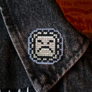 May include: A white embroidered patch with a pixelated design of a face with a frown. The patch is attached to a dark blue denim jacket.