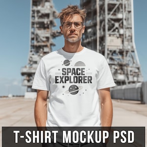 May include: White t-shirt with the words "SPACE EXPLORER" in a bold, black font, accompanied by illustrations of planets and stars. The shirt is worn by a person with glasses, standing in front of a large industrial structure.