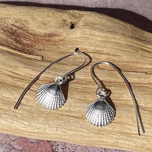 May include: A pair of silver drop earrings featuring small, textured seashell charms. Each earring is suspended from a curved, minimalist silver wire hook that passes through the ear, showcasing a detailed, ribbed fan-shaped shell design.