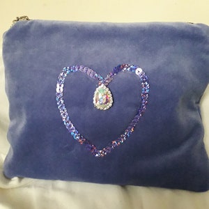 Könnte beinhalten: Eine Clutch aus lila Samt mit einem herzförmigen Design aus schimmernden Pailletten und einem großen, schimmernden Strassstein in der Mitte.
