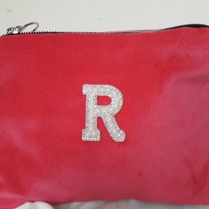 Könnte beinhalten: Eine leuchtend pinkfarbene Samt-Kosmetiktasche mit einem großen weißen Buchstaben "R", der mit weißen Strasssteinen verziert ist.