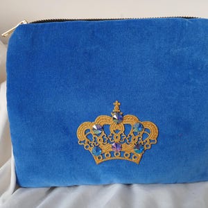 Könnte beinhalten: Eine königlich blaue Samt-Tasche mit einer goldenen, gestickten Krone mit blauen Strasssteinen. Die Tasche hat einen Reißverschluss.