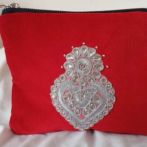 Könnte beinhalten: Eine rote Samttasche mit silbernem Reißverschluss und einem dekorativen Design aus Silber und klaren Edelsteinen. Die verzierte Verzierung ist auf der Vorderseite der Tasche zentriert.