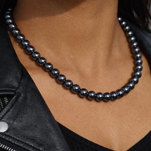 May include: A dark gray beaded necklace. The necklace is composed of small, round beads and is worn against a dark background. The beads have a metallic sheen. The person is wearing a black leather jacket.