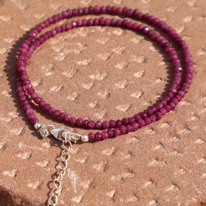 Peut inclure: Un collier délicat composé de deux rangées de petites perles de rubis facettées. Les perles rouge foncé sont enfilées avec un fermoir argenté et une chaîne réglable. Le bijou repose sur une surface texturée brun-rouge.