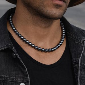 May include: A dark gray beaded necklace. The necklace is composed of numerous small, round beads. The necklace is worn against a black shirt and a dark denim jacket. The beads have a metallic sheen.