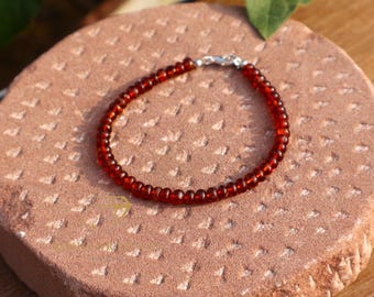 May include: A beaded bracelet featuring small, round, dark red beads. The bracelet is strung on a flexible cord and secured with a silver-colored clasp. The bracelet is resting on a textured, reddish-brown surface.