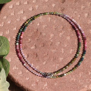 Può includere: Una delicata collana composta da piccole perline colorate nelle tonalità del rosa, verde e viola. La collana è a doppia fila e fissata con una chiusura argentata. Poggia su una superficie strutturata marrone-rossastra.
