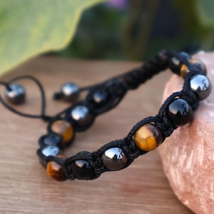 Op de afbeelding: Een zwart macramé armband met een mix van gepolijste kralen. De armband bevat zwarte, zilveren en goudbruine tijgeroogkralen. De verstelbare armband wordt gepresenteerd op een houten oppervlak.