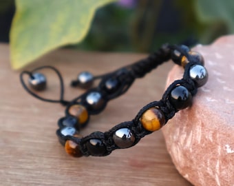 May include: A black macrame bracelet featuring a mix of polished beads. The bracelet includes black, silver, and golden-brown tiger eye beads. The adjustable bracelet is displayed on a wooden surface.