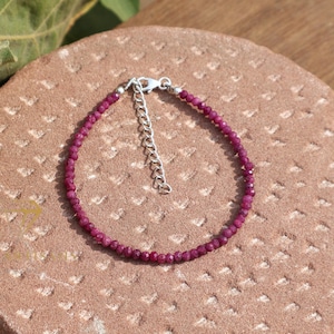 Könnte beinhalten: Ein zartes Armband mit kleinen, facettierten, tiefroten Perlen. Das Armband ist an einer Kette mit einem silberfarbenen Verschluss und Verlängerung befestigt. Der Schmuck wird auf einer strukturierten, rotbraunen Oberfläche präsentiert.