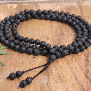 Peut inclure: Un collier de perles en pierre de lave noire avec une fermeture à pompon noué. Le collier est composé de petites perles rondes et texturées. Les perles sont enfilées pour former un long brin. Le collier est présenté sur une surface en bois.