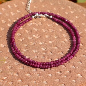 May include: A double-strand necklace featuring small, faceted, deep red gemstones. The necklace has a silver-colored clasp and chain. The gemstones are arranged in a circular pattern on a textured, reddish-brown surface.