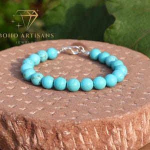Könnte beinhalten: Ein türkisfarbenes Perlenarmband mit Silberverschluss liegt auf einem strukturierten, rotbraunen Stein. Das Armband besteht aus runden, hellblauen Perlen. Das Logo von Boho Artisans Jewelry ist sichtbar.