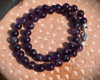 May include: A necklace featuring large, polished, deep purple amethyst beads strung on a purple cord. The necklace is secured with a silver-colored clasp. The beads have a glossy finish and are arranged in a double strand.
