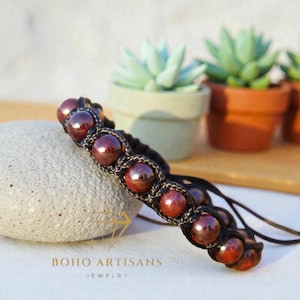 May include: A handmade bracelet featuring dark red, round beads woven into a black macrame design. The bracelet rests on a gray stone, with small potted succulents in the background. The bracelet is a piece of jewelry.