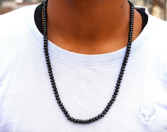 May include: A black beaded necklace with a faceted design. The necklace is worn over a white t-shirt. The beads appear to be small and evenly spaced, creating a simple yet elegant accessory.
