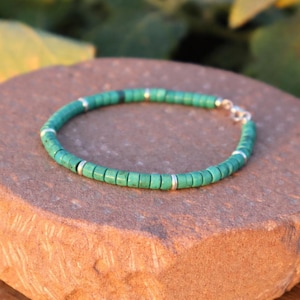May include: A green beaded bracelet with silver detailing. The bracelet features small, cylindrical green beads interspersed with silver-coloured metal spacers. The bracelet is shown on a brown, textured surface.