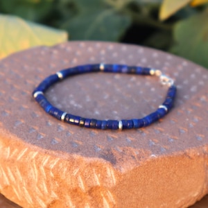 May include: A dark blue beaded bracelet with silver-coloured accents. The bracelet features small, dark blue beads and silver-coloured beads. It rests on a textured, reddish-brown surface. The clasp is silver.