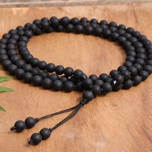 May include: A black matte beaded necklace with a tassel closure. The necklace is made of small, round beads and is displayed on a wooden surface. A green leafy plant is in the corner.