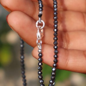 May include: A close-up of a black beaded necklace with a silver clasp. The necklace is made of small, faceted beads that reflect light. The clasp is a simple, secure design.