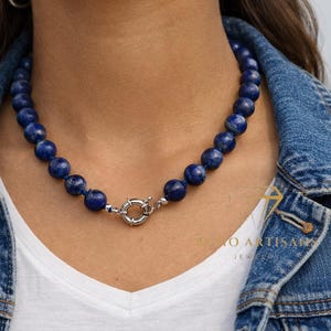 May include: A beaded necklace featuring dark blue, spherical beads. The necklace is secured with a silver-colored clasp. The person is wearing a white shirt and a denim jacket.