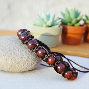 May include: A bracelet featuring a series of dark red, polished spherical beads secured within a black macrame cord design. The bracelet includes adjustable cord ends with small matching beads, showcasing a detailed knotted pattern throughout.