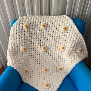 May include: White crocheted blanket with yellow daisy flowers. The blanket is folded and placed on a blue armchair.