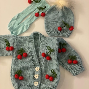 May include: A light blue knitted cardigan with a cherry motif, featuring a button closure and long sleeves. The cardigan is paired with a matching light blue knitted hat with a white pom-pom and a pair of light blue knitted socks with cherry embellishments.
