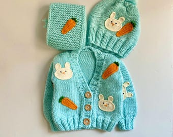 May include: A light blue knitted baby cardigan with a bunny and carrot design. The set includes a matching hat and scarf.