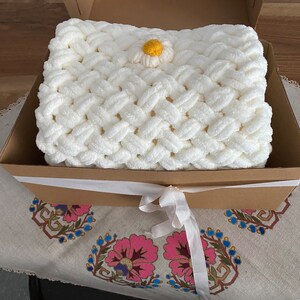 May include: A white crocheted blanket with a yellow and white daisy flower accent, presented in a brown gift box with a white ribbon.