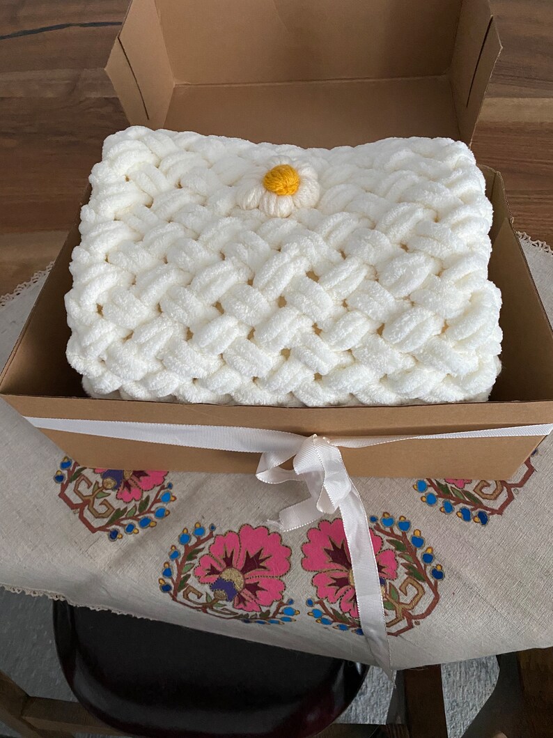 May include: A white crocheted blanket with a yellow and white daisy flower accent, presented in a brown gift box with a white ribbon.