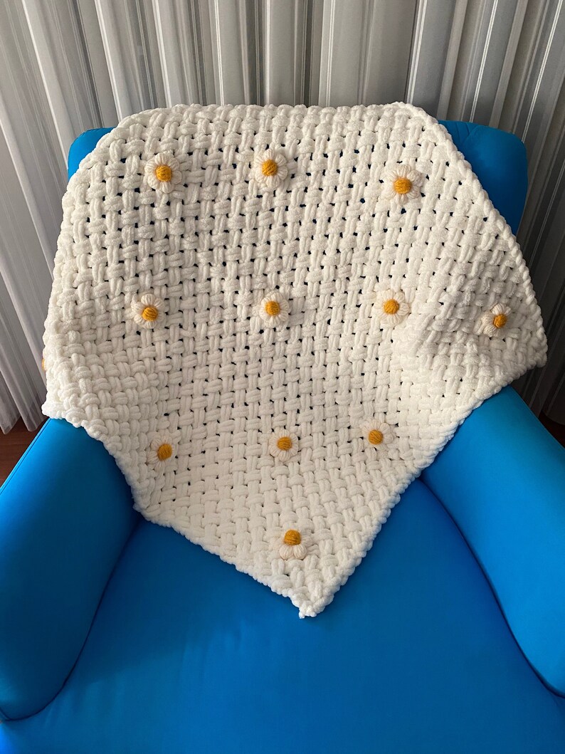 May include: White crocheted blanket with yellow daisy flowers. The blanket is folded and placed on a blue armchair.