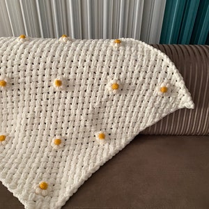May include: A white crocheted throw blanket with yellow daisy-shaped embellishments. The blanket is folded and draped over a brown couch.