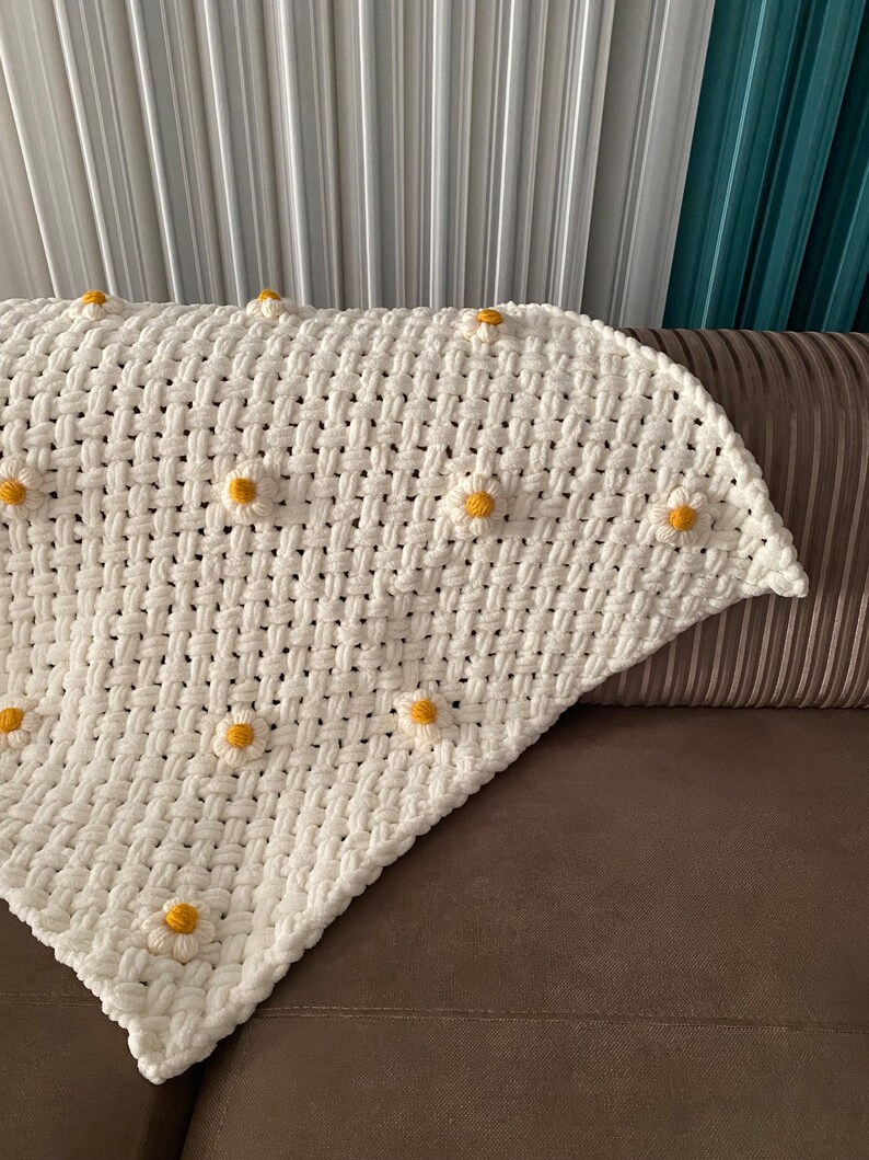 May include: A white crocheted throw blanket with yellow daisy-shaped embellishments. The blanket is folded and draped over a brown couch.