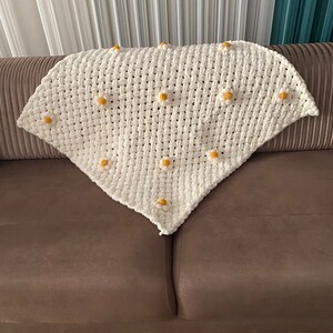 May include: A white crocheted blanket with a triangular shape and yellow daisy accents.