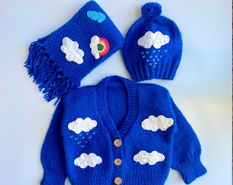 May include: A blue knitted cardigan with white cloud appliques and a rainbow detail. The cardigan has a v-neck and button closure. A matching blue knitted hat with a white cloud applique and a pom-pom on top is also included. A blue knitted scarf with white cloud appliques and a rainbow detail is also included.