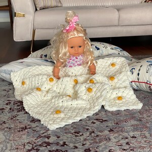 May include: A white crocheted blanket with yellow daisy flowers. A doll with blonde hair and a pink bow is sitting on the blanket.