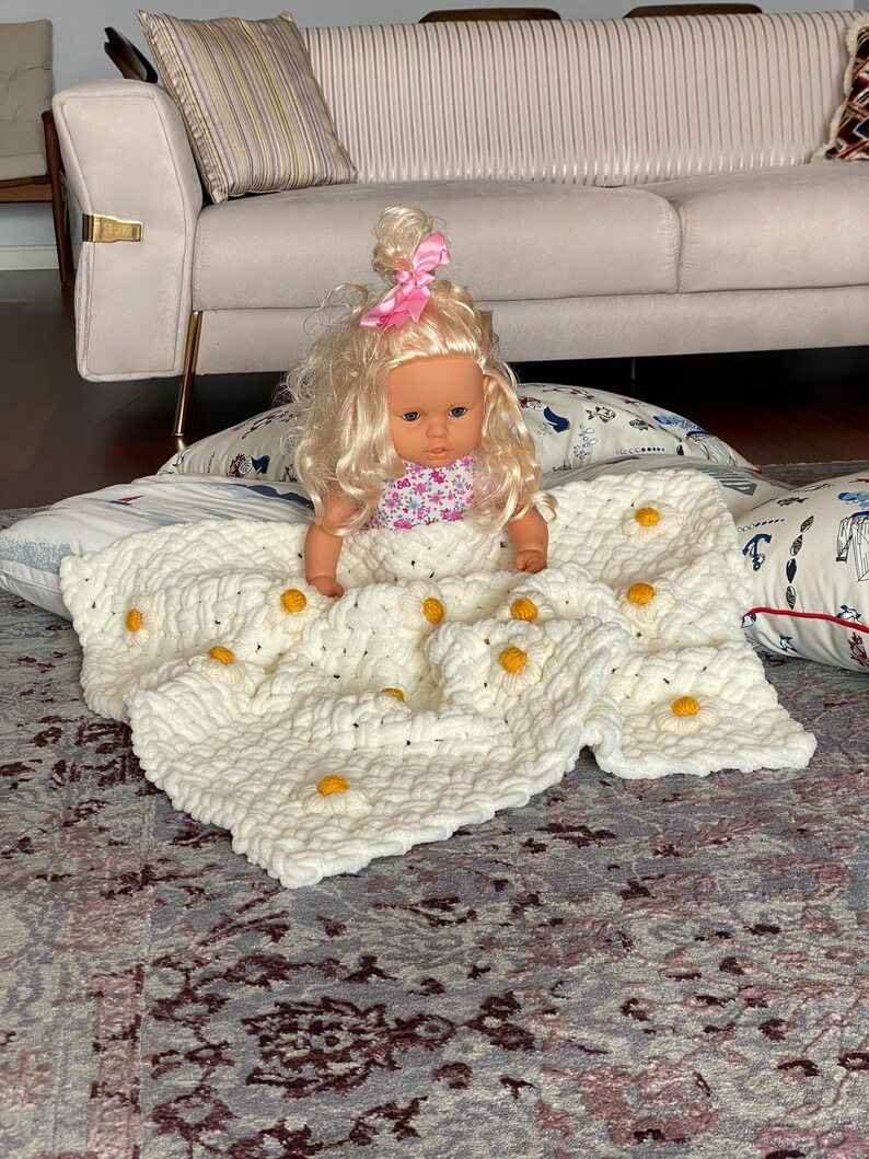 May include: A white crocheted blanket with yellow daisy flowers. A doll with blonde hair and a pink bow is sitting on the blanket.